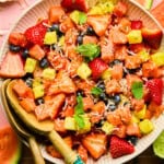 A bowl of vibrant Watermelon Fruit Salad with strawberries, pineapple, blueberries, shredded coconut, and mint leaves. Wooden serving utensils rest in the bowl, and fruit slices decorate the pink tiled surface underneath.