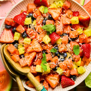 A bowl of vibrant Watermelon Fruit Salad with strawberries, pineapple, blueberries, shredded coconut, and mint leaves. Wooden serving utensils rest in the bowl, and fruit slices decorate the pink tiled surface underneath.