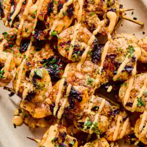 BBQ Prawn Skewers grilled to perfection, topped with a creamy, orange-colored sauce and garnished with chopped herbs, served on a light-colored oval plate.