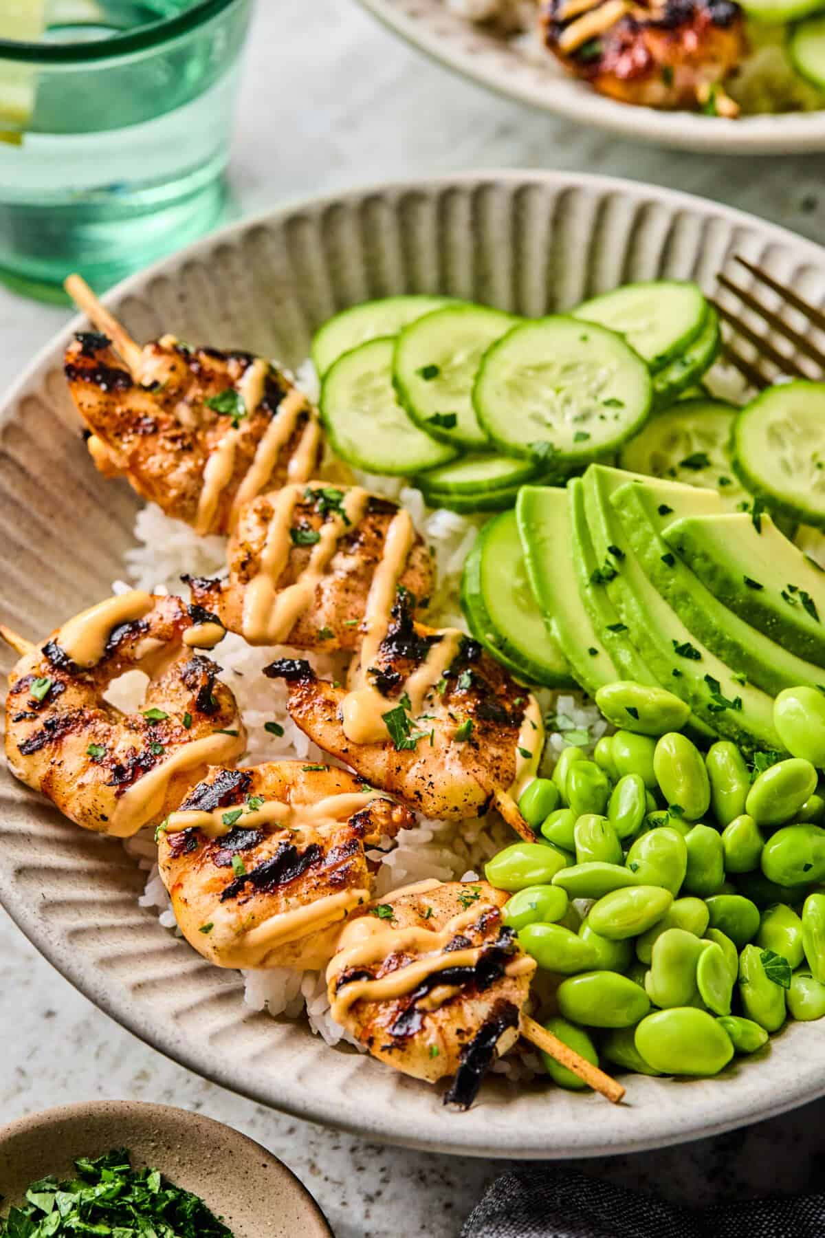 A bowl filled with BBQ prawn skewers drizzled with sauce, white rice, sliced cucumber, avocado, and edamame, garnished with herbs. A glass of water and a small dish of chopped herbs are nearby.