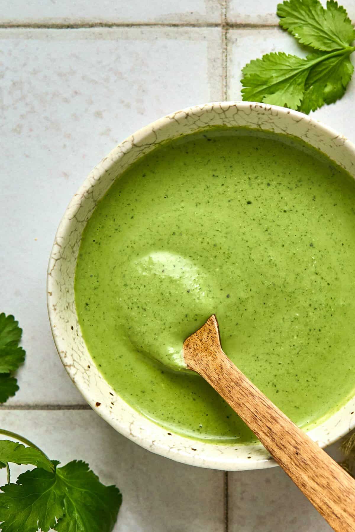 A ceramic bowl brimming with creamy cilantro dressing sits alongside a wooden spoon. Fresh green herbs are scattered on the white tiled surface, enhancing the dishs vibrant appeal.