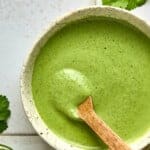 A ceramic bowl brimming with creamy cilantro dressing sits alongside a wooden spoon. Fresh green herbs are scattered on the white tiled surface, enhancing the dishs vibrant appeal.