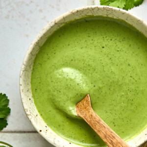 A ceramic bowl brimming with creamy cilantro dressing sits alongside a wooden spoon. Fresh green herbs are scattered on the white tiled surface, enhancing the dishs vibrant appeal.