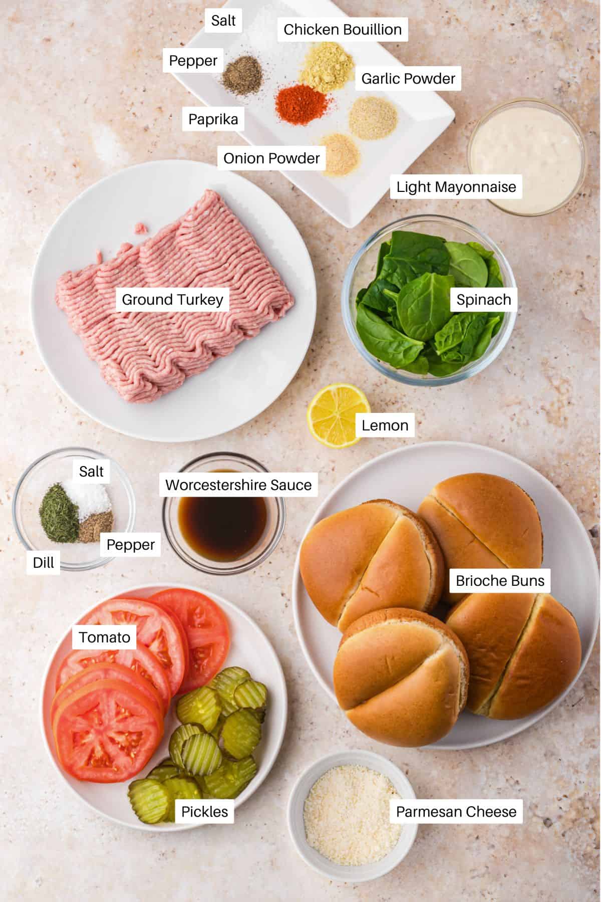 Ingredients for air fryer turkey burgers are arranged on a white surface: ground turkey, brioche buns, spinach, tomato, pickles, lemon, parmesan cheese, mayonnaise, seasonings, Worcestershire sauce, and dill. Labels identify each item.