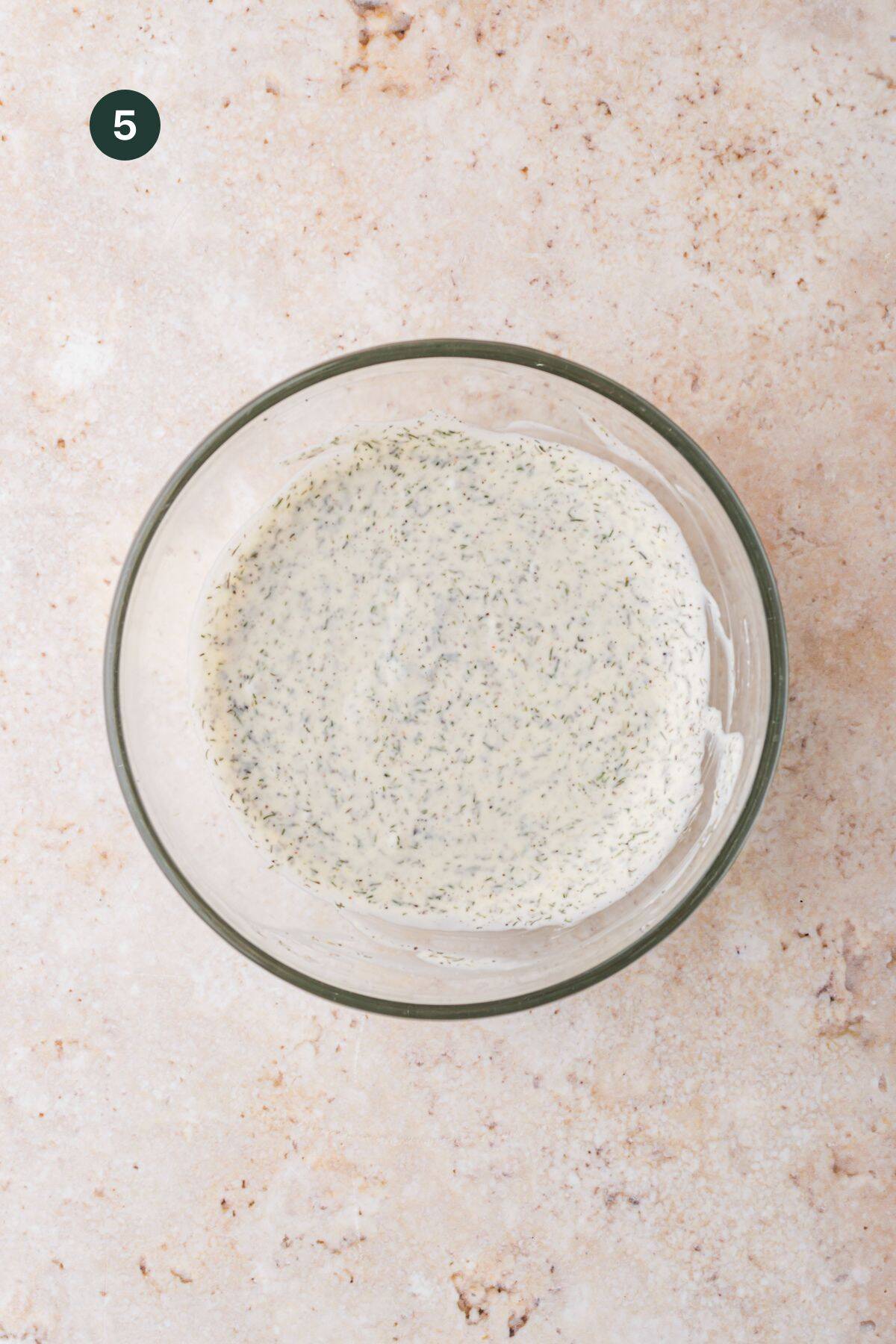 A glass bowl filled with creamy, speckled ranch dressing sits on a light marble surface—perfect for dipping air fryer turkey burgers. A dark green circle with the number 5 is in the upper left corner.