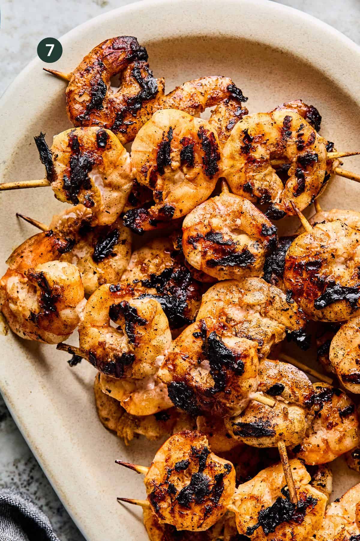 BBQ Prawn Skewers with visible char marks are arranged on a beige oval plate. The shrimp appear seasoned and cooked, with a slightly smoky, crispy exterior.