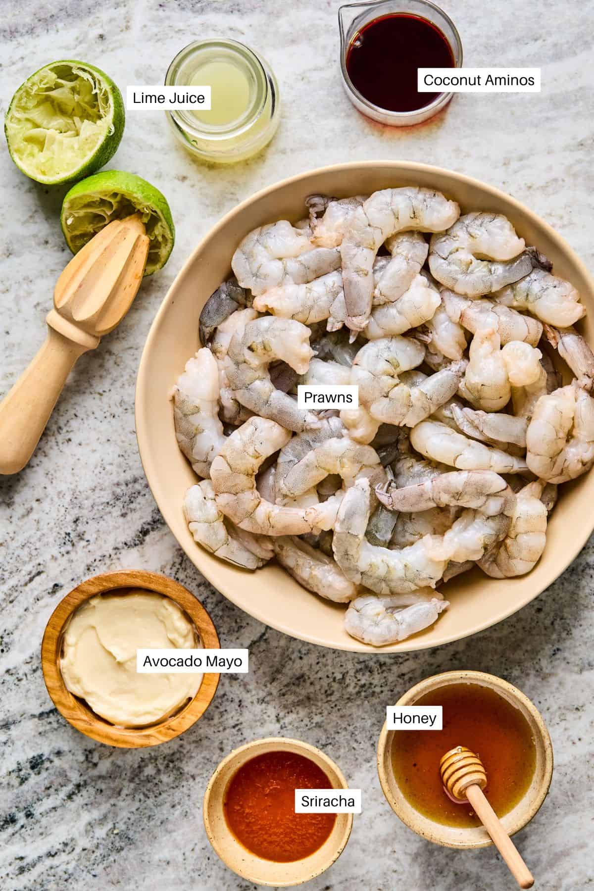 A bowl of raw prawns for BBQ Prawn Skewers is surrounded by labeled ingredients: lime juice, coconut aminos, avocado mayo, sriracha, and honey on a marble surface. A juiced lime and wooden juicer are also visible.