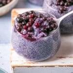 A glass jar brimming with purple chia pudding is crowned with blueberries, reminiscent of blueberry overnight oats. A spoon gracefully lifts a portion dotted with berries. Other jars and a bowl of blueberries peek from the background, all resting on a light-colored wooden board.