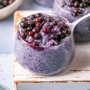 A glass jar brimming with purple chia pudding is crowned with blueberries, reminiscent of blueberry overnight oats. A spoon gracefully lifts a portion dotted with berries. Other jars and a bowl of blueberries peek from the background, all resting on a light-colored wooden board.