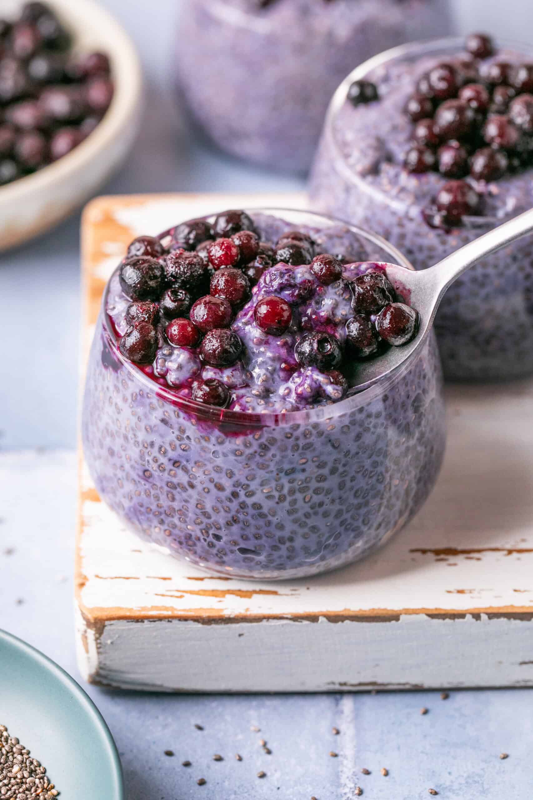 A glass jar brimming with purple chia pudding is crowned with blueberries, reminiscent of blueberry overnight oats. A spoon gracefully lifts a portion dotted with berries. Other jars and a bowl of blueberries peek from the background, all resting on a light-colored wooden board.