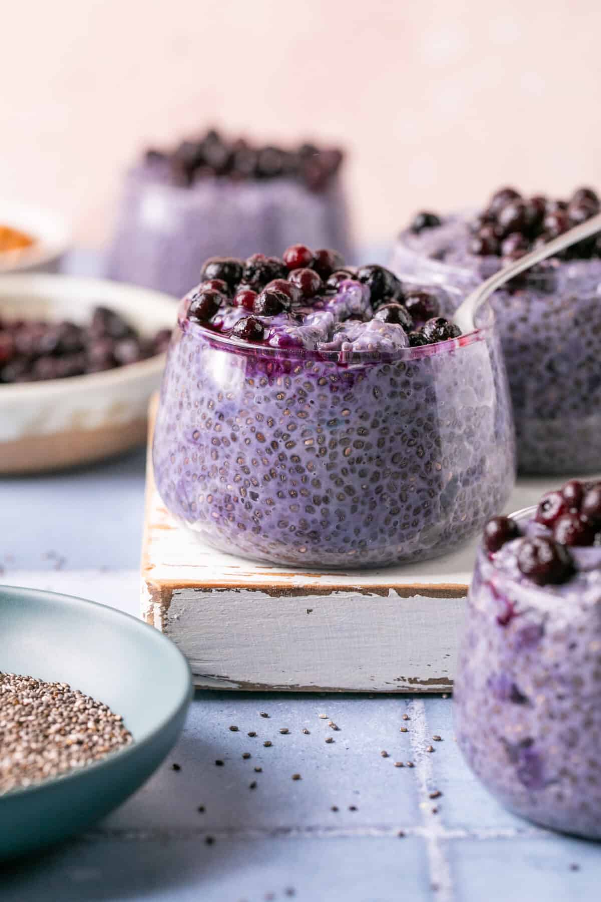 Glass cups filled with delicious blueberry chia pudding, reminiscent of blueberry overnight oats, are topped with fresh blueberries on a white wooden platform. A spoon rests in one cup, while dishes with extra blueberries and chia seeds complete the scene on a light blue surface.