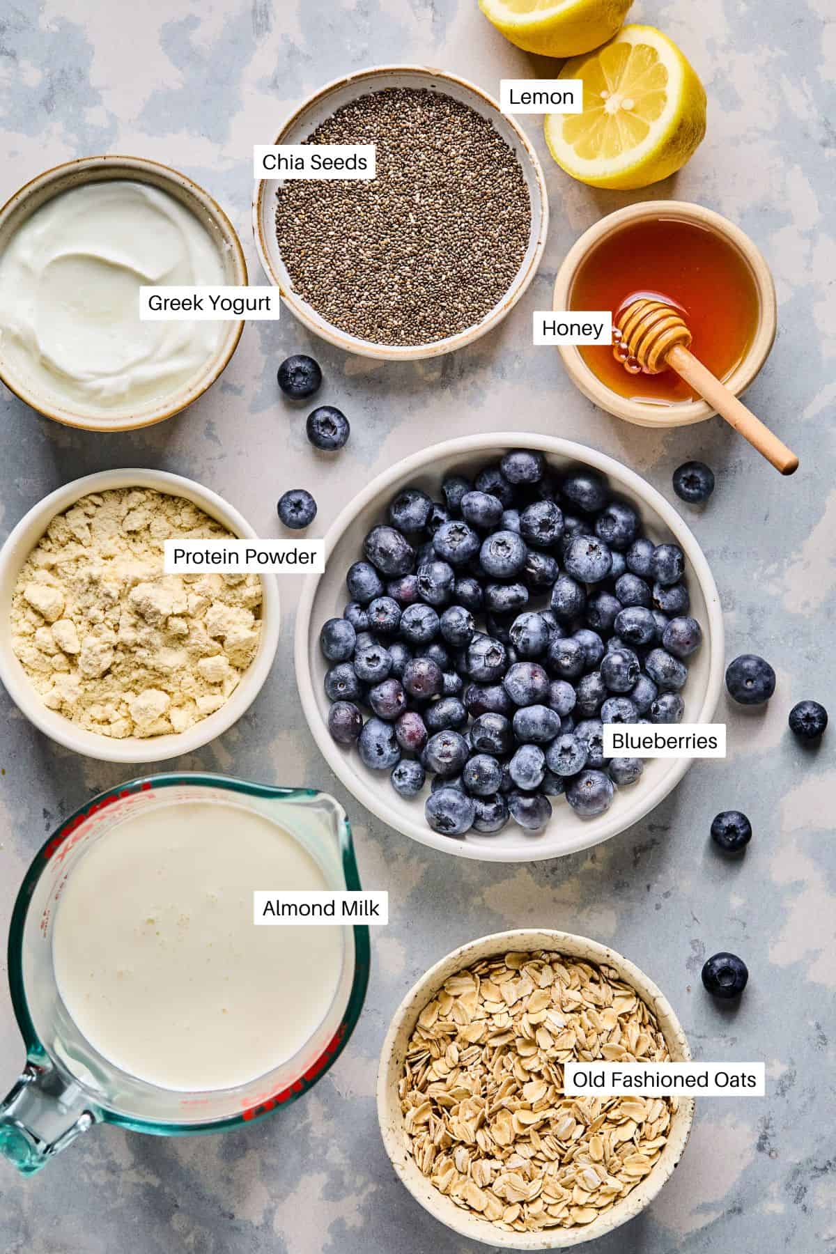 Top view of smoothie ingredients labeled: chia seeds, lemon, Greek yogurt, honey, protein powder, frozen wild blueberries, almond milk, and old fashioned oats. Lemon halves and a honey dipper are also visibleโperfect for creating blueberry overnight oats with a zesty twist.