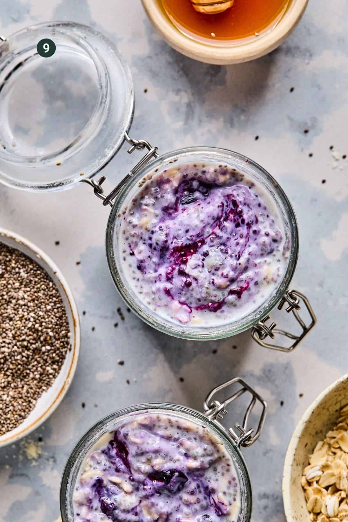Top view of two glass jars filled with blueberry overnight oats, topped with berries. Surrounding them are chia seeds, a glass of water, oats, and a bowl of honey. The table has a light, textured surface.