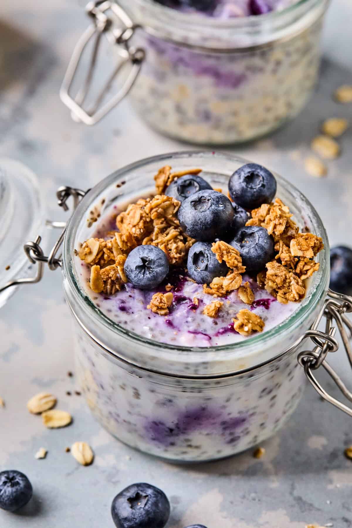 A jar of blueberry overnight oats topped with blueberries and granola. The oats have a marbled effect with swirls of blueberry throughout. The jar is sealed with a metal clasp lid, and the background has scattered oats and more blueberries.