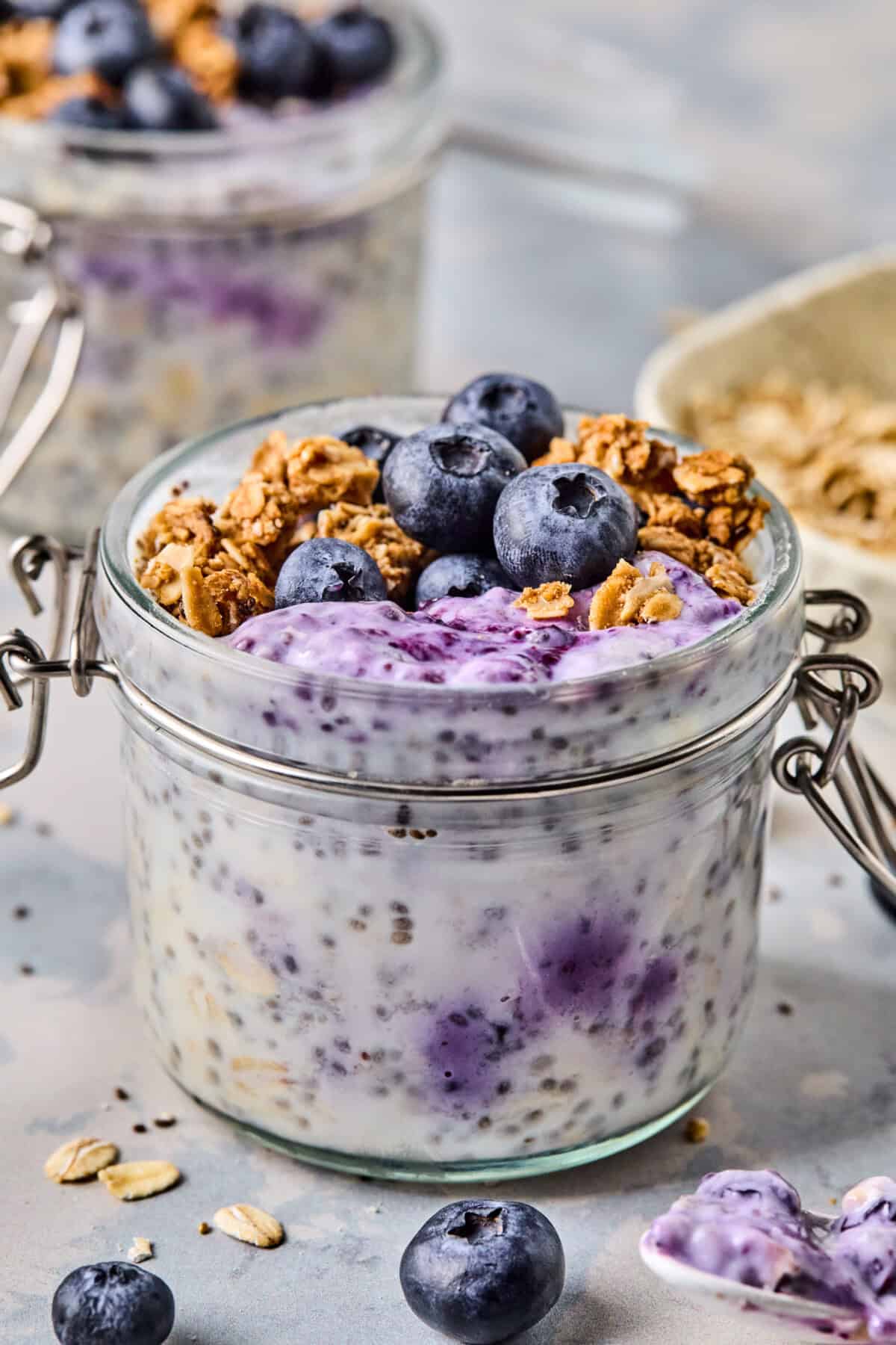 A glass jar brimming with blueberry overnight oats is topped with fresh blueberries, granola, and chia seeds. The oat mixture swirls with a vibrant purple hue from mixed berries. In the background, a spoon and more oats eagerly await their turn to be enjoyed.