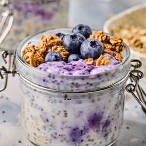 A glass jar brimming with blueberry overnight oats is topped with fresh blueberries, granola, and chia seeds. The oat mixture swirls with a vibrant purple hue from mixed berries. In the background, a spoon and more oats eagerly await their turn to be enjoyed.