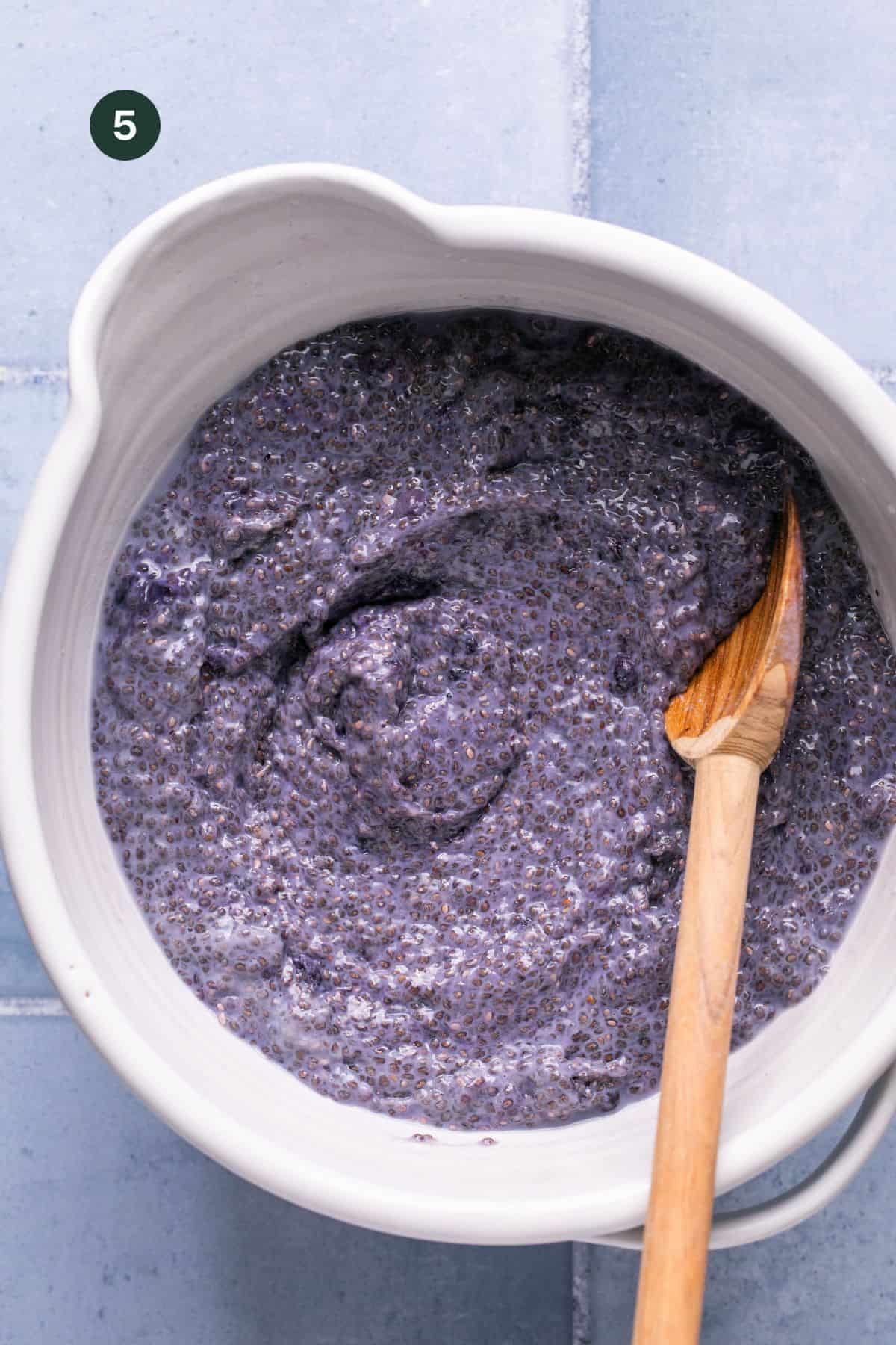 A white bowl filled with purple chia seed pudding, reminiscent of blueberry overnight oats, sits on a blue tiled surface. A wooden spoon rests inside the bowl. The number five is in a dark circular area in the top left corner.