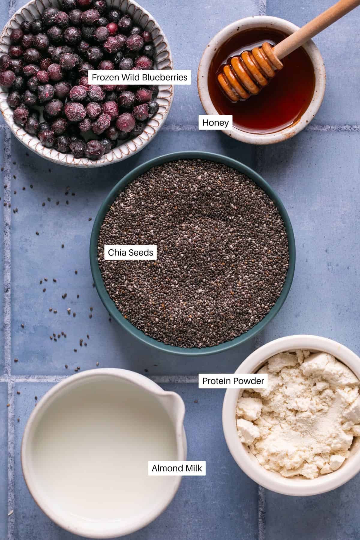 A top-down view of ingredients for blueberry overnight oats on a blue tile surface: a bowl of frozen wild blueberries, a small dish of honey with a honey dipper, chia seeds, protein powder, and a small jug of almond milk.
