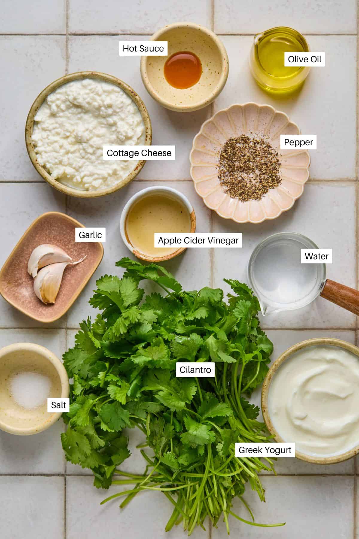 An overhead view of various ingredients labeled: cottage cheese, olive oil, pepper, hot sauce, apple cider vinegar, water, Greek yogurt, cilantro for a creamy cilantro dressing, garlic, and salt. All are arranged on a white tile surface.