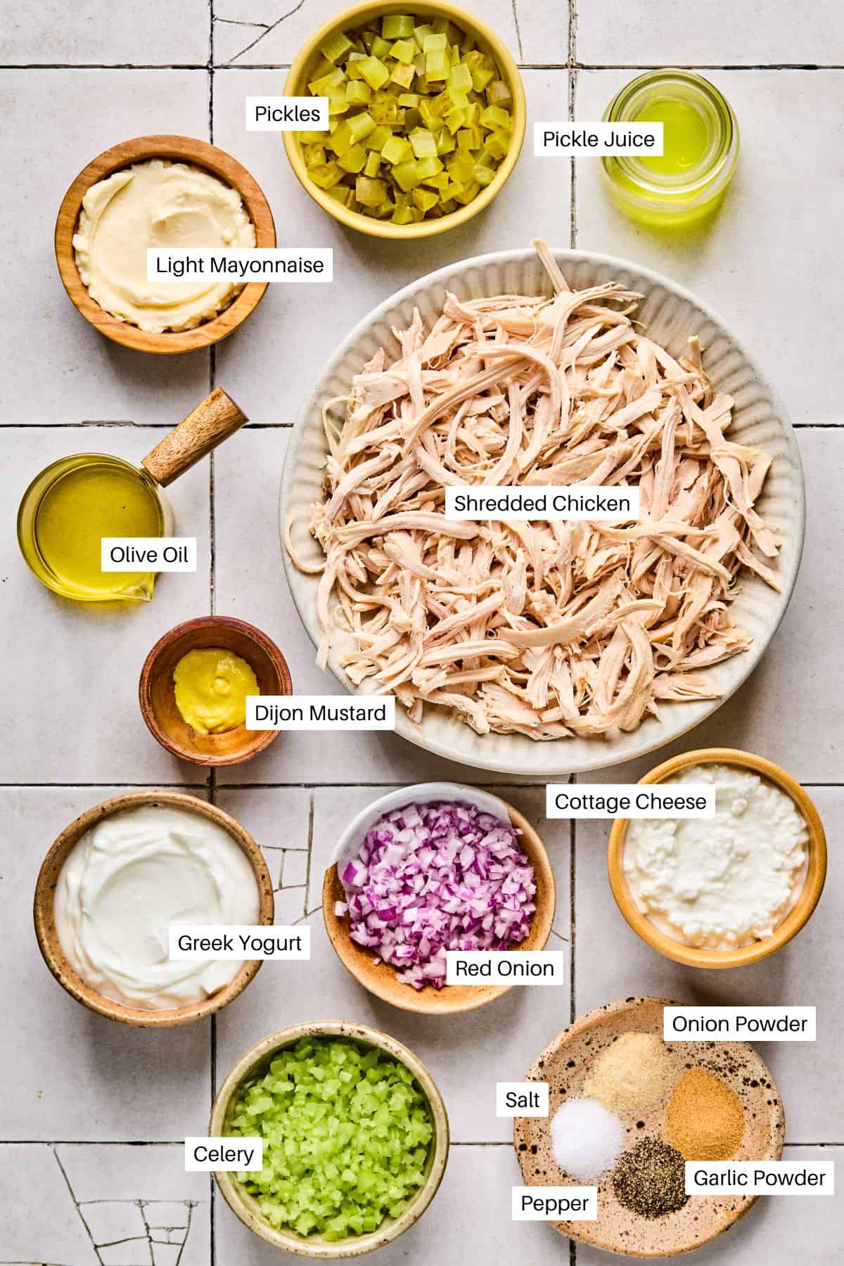 A top-down view of bowls with ingredients for Dill Pickle Chicken Salad: shredded chicken, cottage cheese, Greek yogurt, celery, red onion, Dijon mustard, light mayonnaise, olive oil, pickles and pickle juice, pepper, onion powder, and garlic powder.