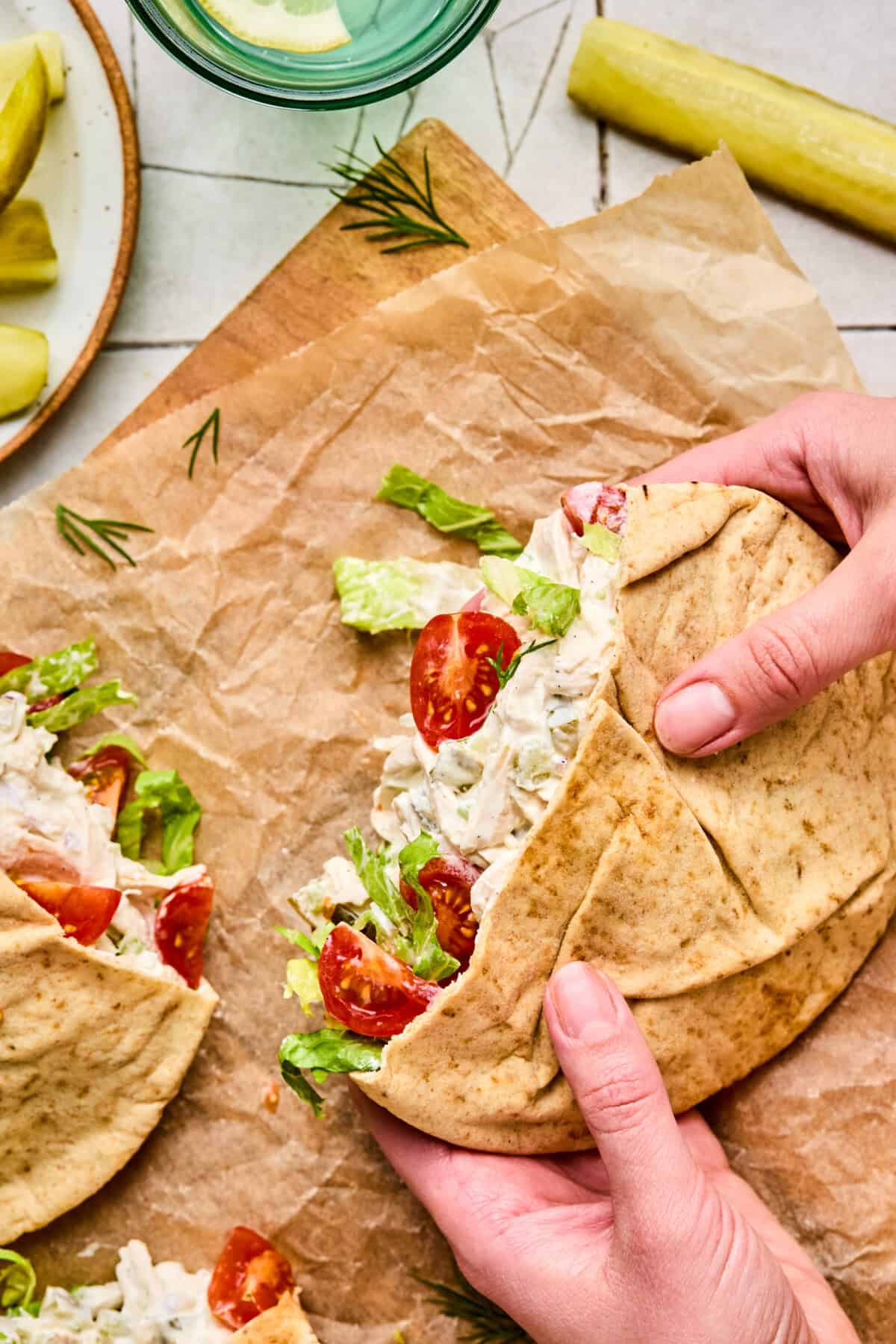 A person holds a pita bread filled with Dill Pickle Chicken Salad, lettuce, and tomato. Another stuffed pita sits nearby on parchment paper, with pickles and a glass of water in the background.