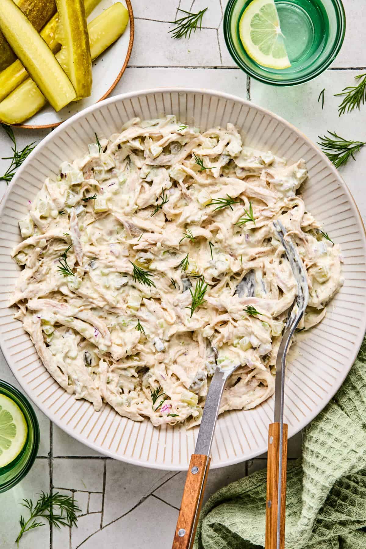 A bowl of creamy Dill Pickle Chicken Salad garnished with fresh dill, with metal tongs resting inside. Nearby, sliced pickles are on a plate, and a glass of water with lemon sits beside a green cloth.
