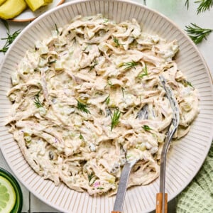 A bowl of creamy Dill Pickle Chicken Salad garnished with fresh dill, with metal tongs resting inside. Nearby, sliced pickles are on a plate, and a glass of water with lemon sits beside a green cloth.