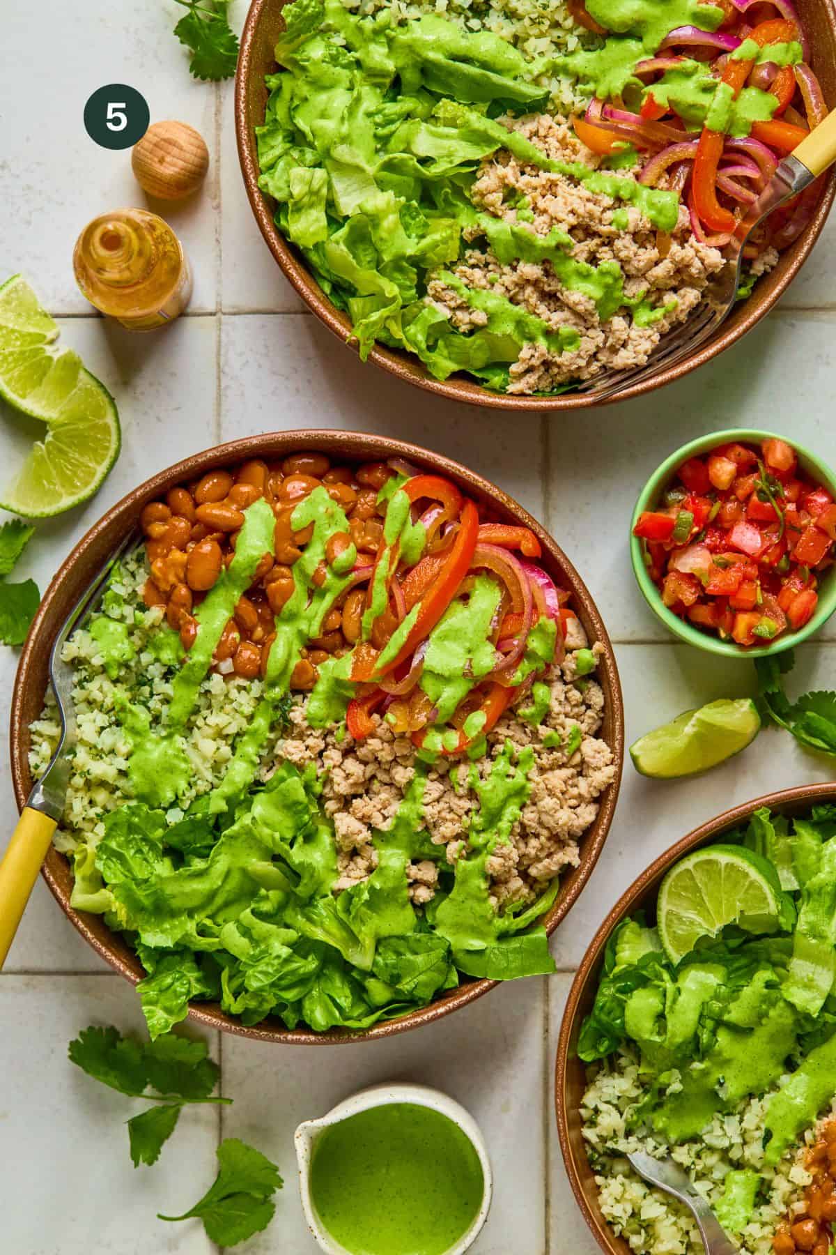 Three bowls of vibrant salad with lettuce, chopped veggies, beans, and cilantro, reminiscent of a recipe for taco bowl, are drizzled with green dressing. Lime wedges and a small bowl of salsa are on the side. A small bottle and nut rest nearby on a white tiled surface.