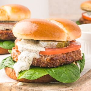 A close-up of an air fryer turkey burger on a wooden board, topped with spinach, tomato slices, pickles, and creamy white sauce in a shiny brioche bun. Another burger is blurred in the background.