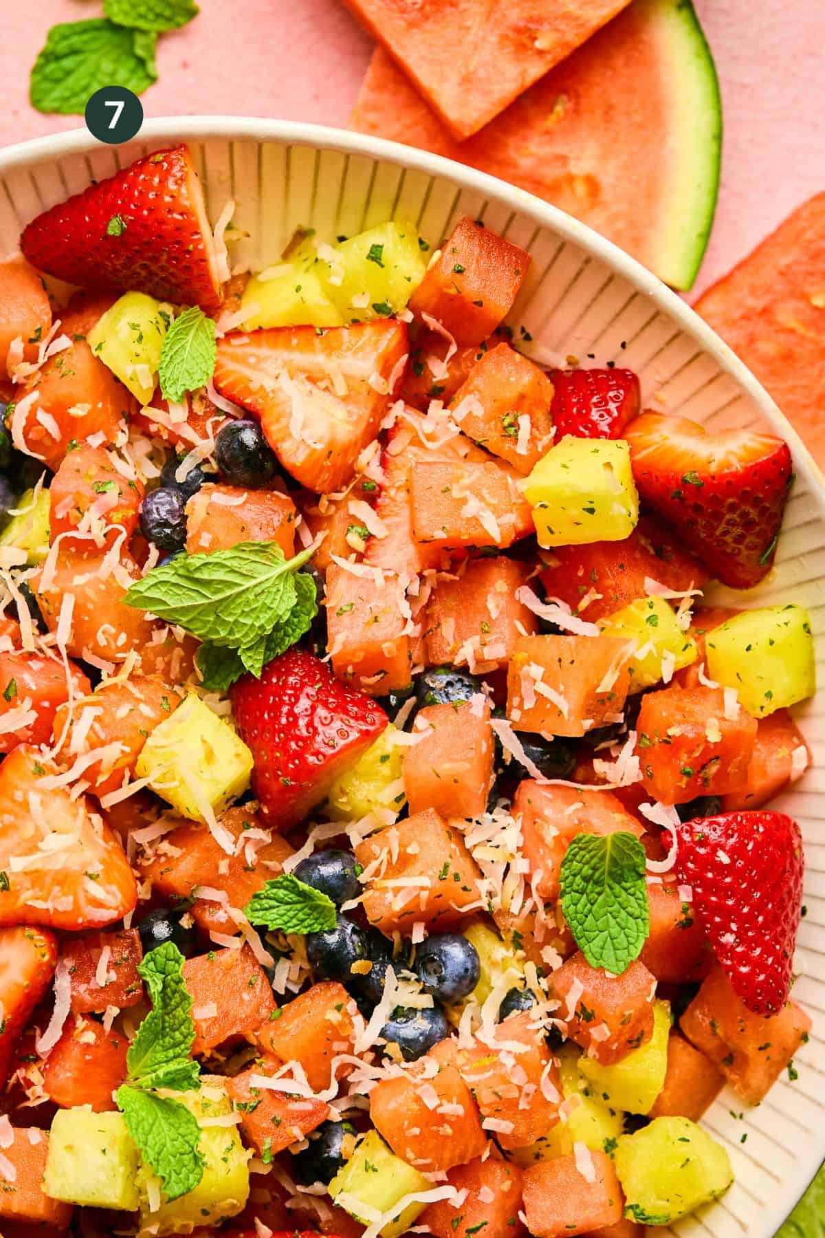 A close-up of a Watermelon Fruit Salad in a white bowl, featuring diced watermelon, pineapple, strawberries, blueberries, shredded coconut, and fresh mint leaves, with slices of watermelon in the background.