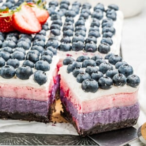 A layered Berry Cake bar with a chocolate crust, purple, pink, and white cream layers, topped with rows of fresh blueberries and sliced strawberries. A square piece is cut out to reveal the vibrant layers inside.