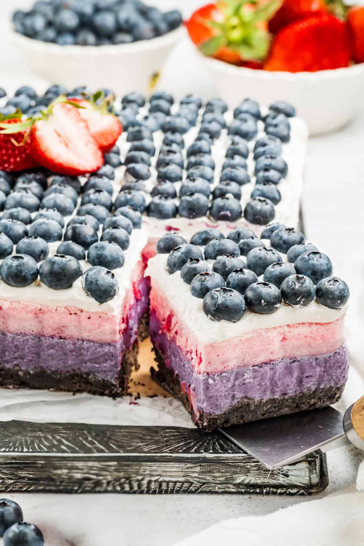 A layered Berry Cake bar with a chocolate crust, purple, pink, and white cream layers, topped with rows of fresh blueberries and sliced strawberries. A square piece is cut out to reveal the vibrant layers inside.