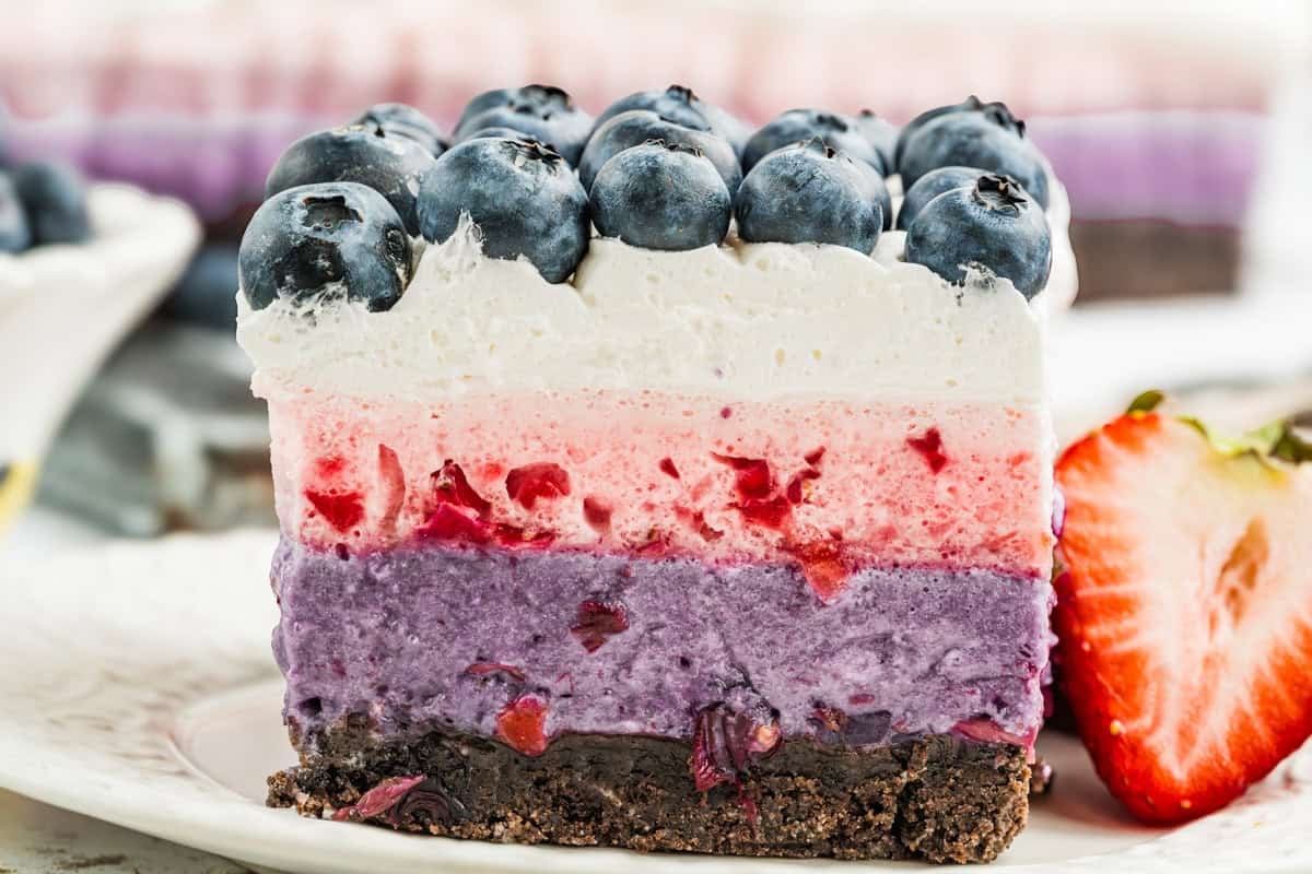 A close-up of a layered Berry Cake with a crumbly base, purple berry layer, pink layer, white cream layer, and topped with fresh blueberries, served on a plate with a sliced strawberry.