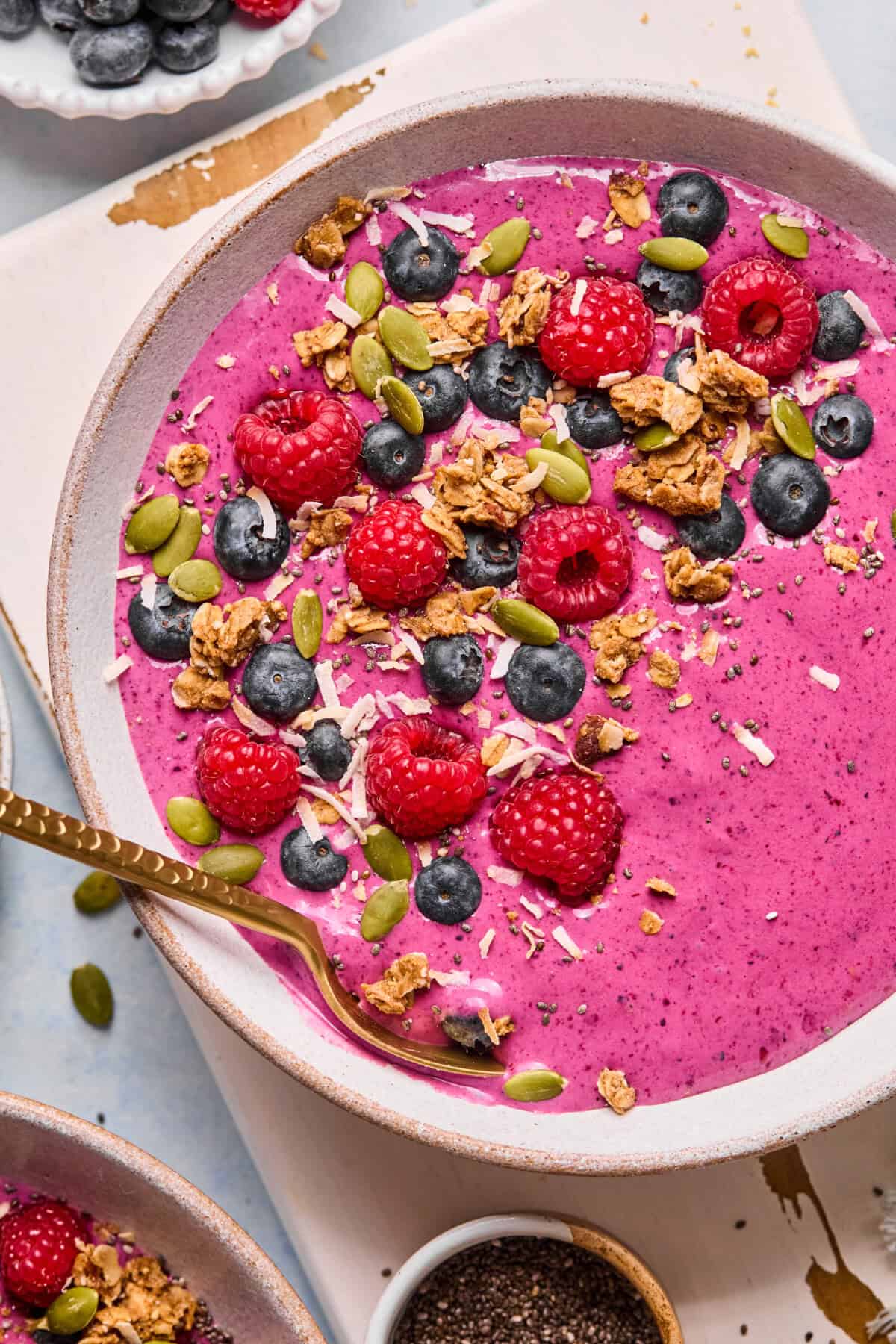 A vibrant berry smoothie bowl topped with fresh raspberries, blueberries, granola, pumpkin seeds, shredded coconut, and chia seeds. A gold spoon rests in the bowl. The colors are bright and appetizing.