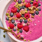 A vibrant berry smoothie bowl topped with fresh raspberries, blueberries, granola, pumpkin seeds, shredded coconut, and chia seeds. A gold spoon rests in the bowl. The colors are bright and appetizing.