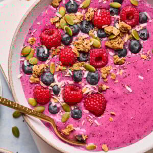 A vibrant berry smoothie bowl topped with fresh raspberries, blueberries, granola, pumpkin seeds, shredded coconut, and chia seeds. A gold spoon rests in the bowl. The colors are bright and appetizing.