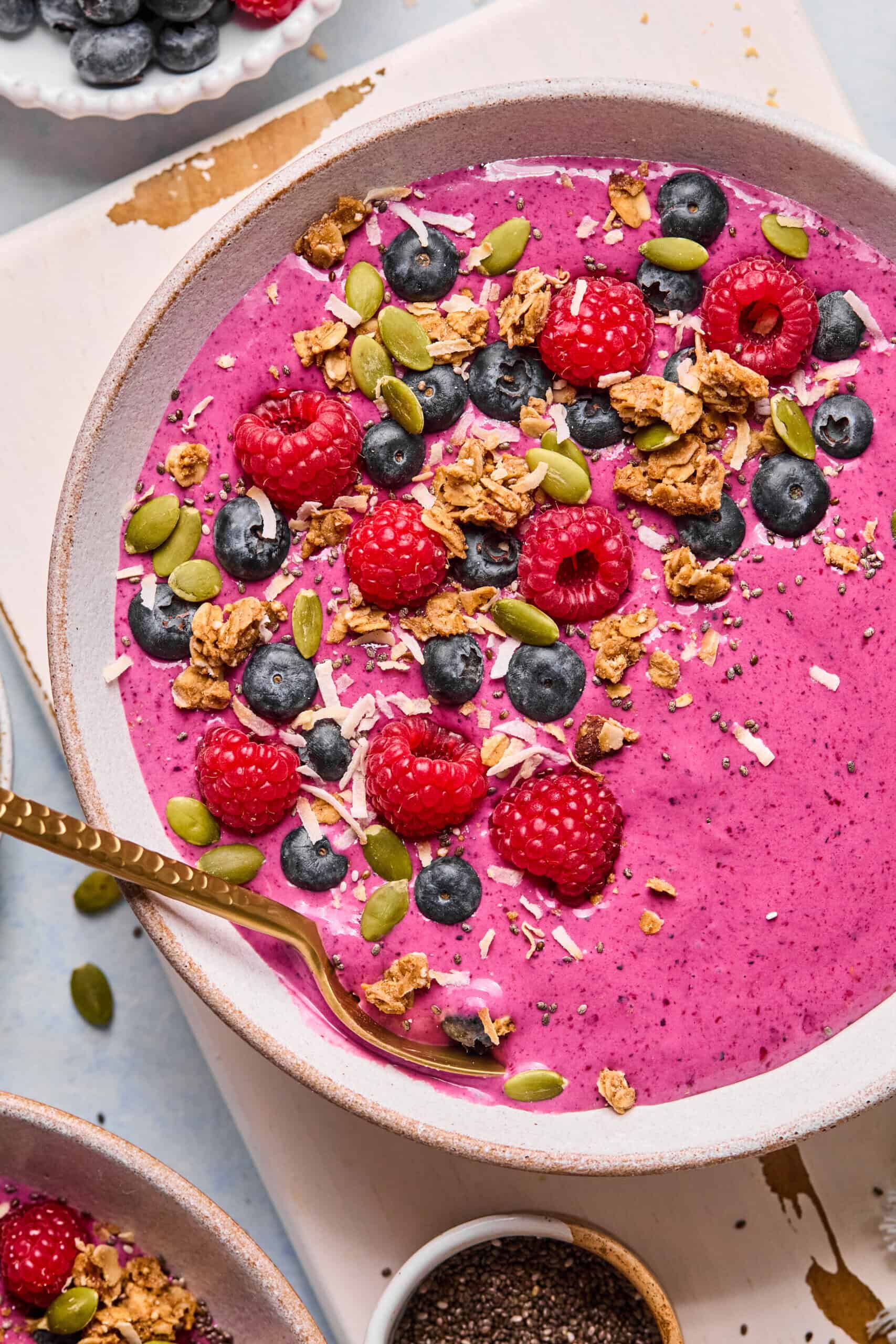 A vibrant berry smoothie bowl topped with fresh raspberries, blueberries, granola, pumpkin seeds, shredded coconut, and chia seeds. A gold spoon rests in the bowl. The colors are bright and appetizing.