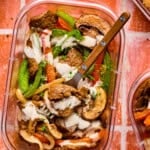 A clear container holds a vibrant mix of sliced beef, green bell peppers, onions, mushrooms, and red peppers drizzled with a creamy sauce—classic Philly cheesesteak bowl flavors. A fork rests inside on a reddish tile surface.
