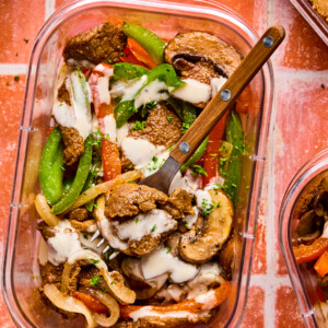 A clear container holds a vibrant mix of sliced beef, green bell peppers, onions, mushrooms, and red peppers drizzled with a creamy sauceโclassic Philly cheesesteak bowl flavors. A fork rests inside on a reddish tile surface.