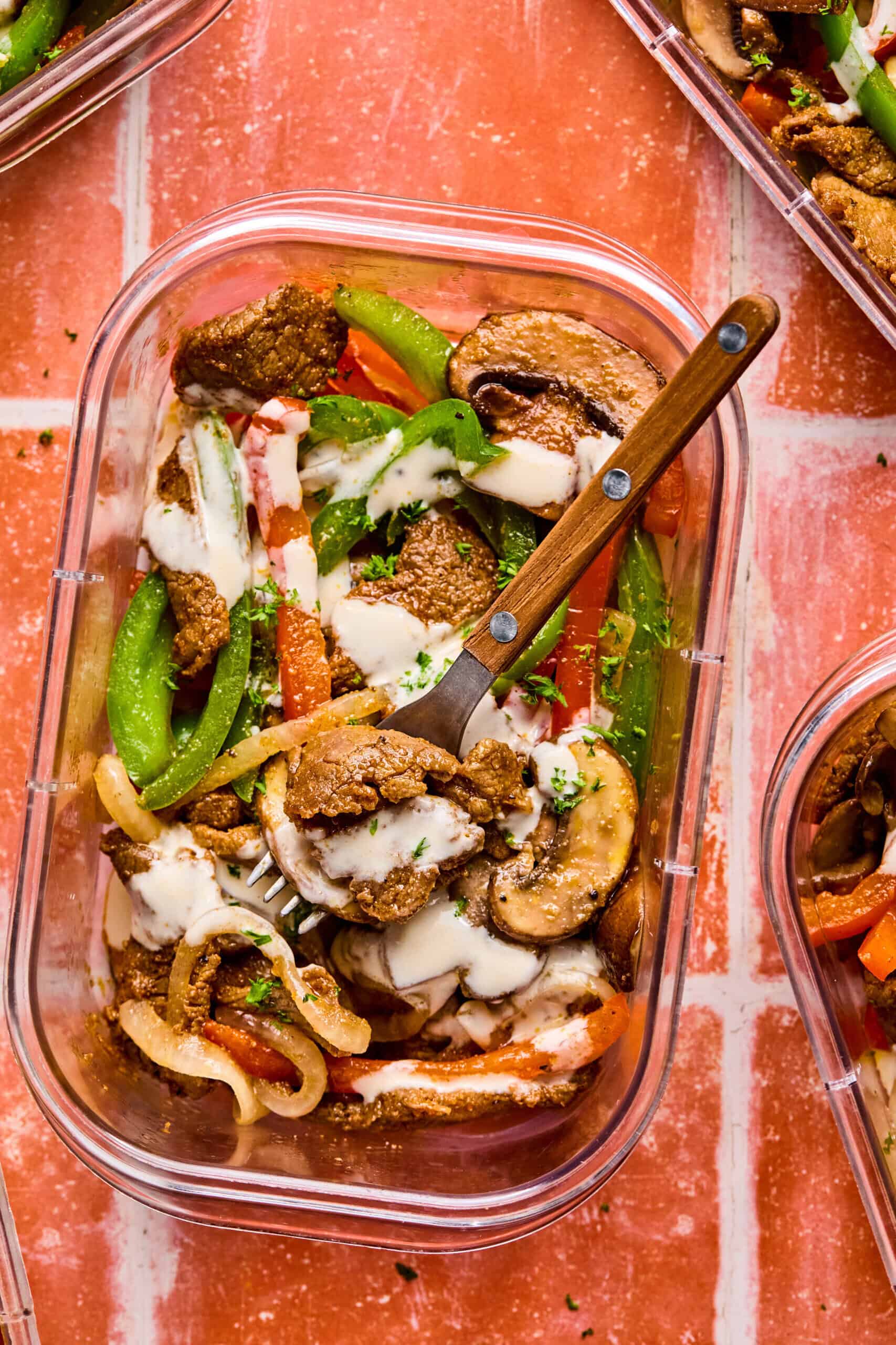 A clear container holds a vibrant mix of sliced beef, green bell peppers, onions, mushrooms, and red peppers drizzled with a creamy sauce—classic Philly cheesesteak bowl flavors. A fork rests inside on a reddish tile surface.