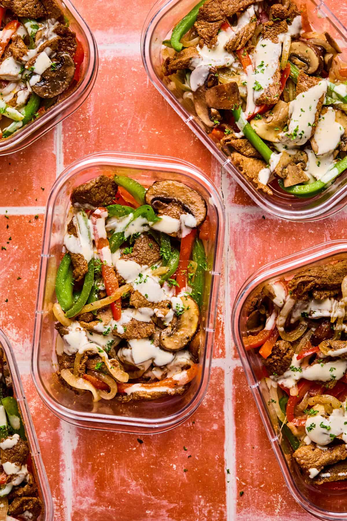 Five clear meal prep containers filled with Philly cheesesteak bowlsโsautรฉed beef strips, bell peppers, onions, mushrooms, and creamy white sauceโare arranged on an orange tiled surface. Fresh herbs are sprinkled on top.