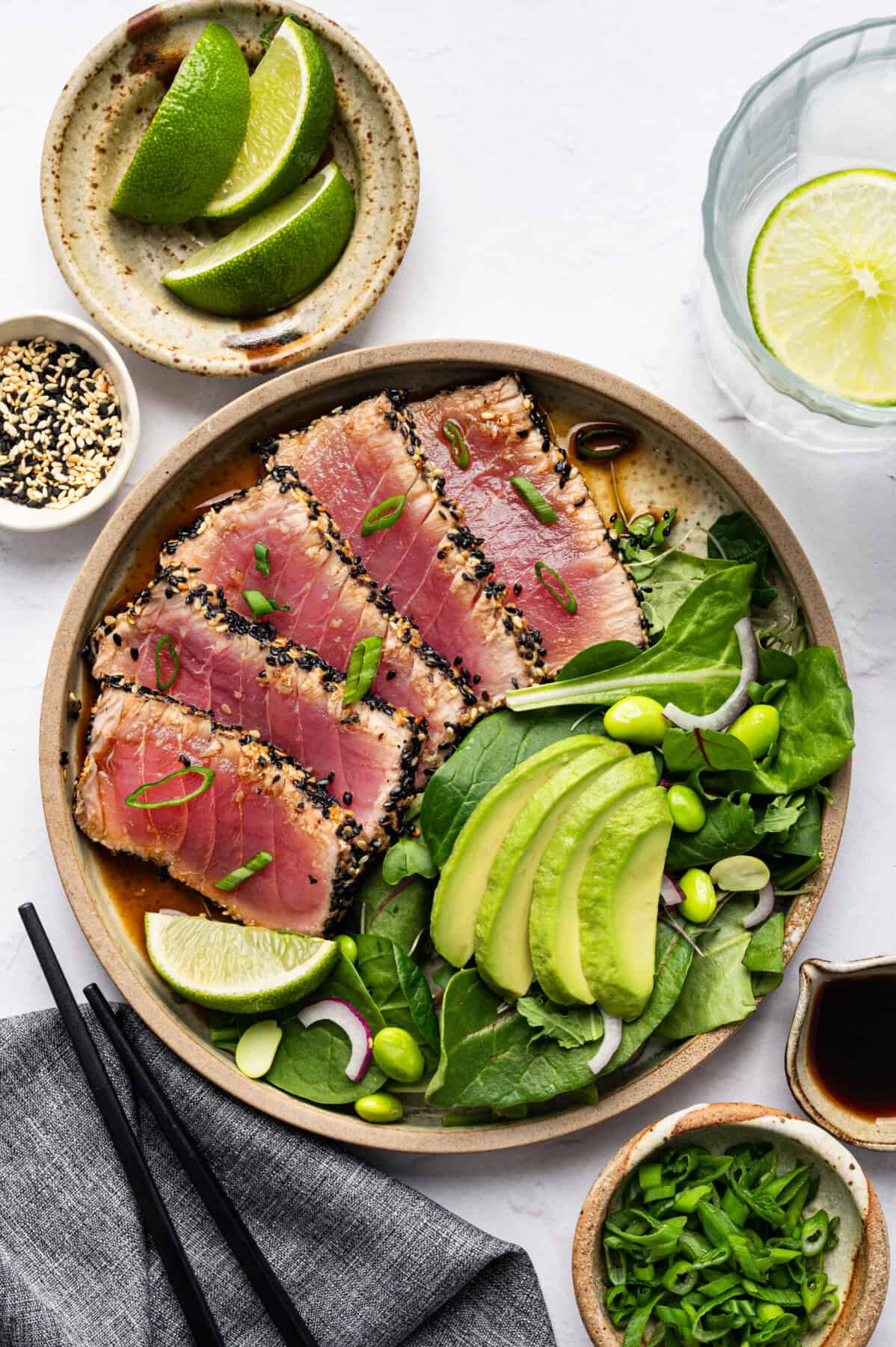 A bowl with sesame-crusted seared tuna slices, fresh spinach, avocado, edamame, and red onion joins baked chicken cutlets and a lime wedge. Served with soy sauce, sesame seeds, lime on the sideโchopsticks and a glass of water nearby.