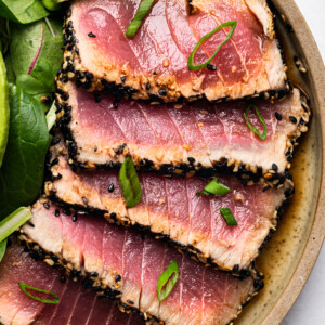 Five slices of seared ahi tuna with a sesame seed crust, topped with green onions, arranged on a plate with spinach leaves, red onion, and a lime wedgeโan elegant alternative to classic baked chicken cutlets. The tuna is pink and lightly seared on the edges.