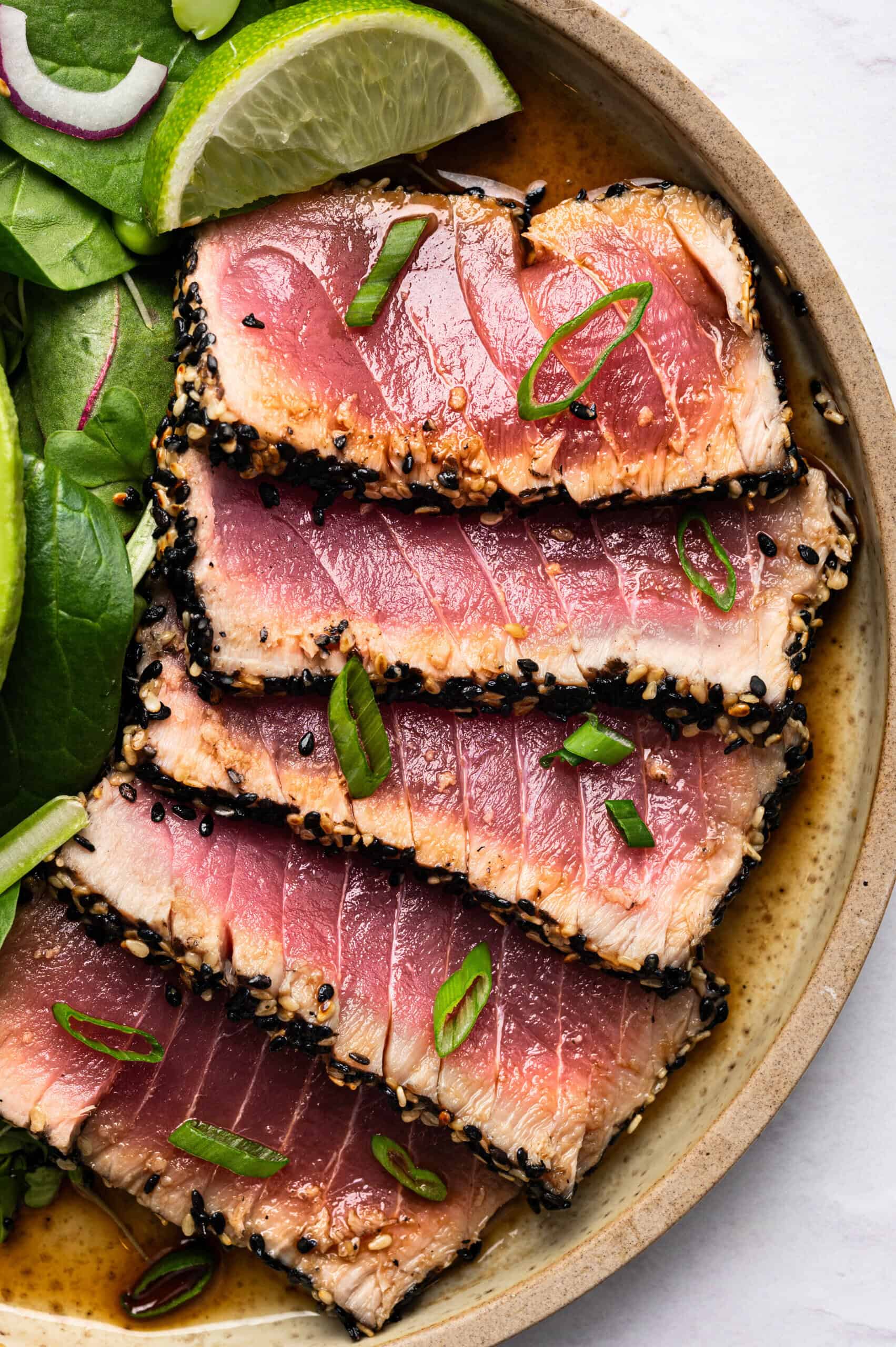 Five slices of seared ahi tuna with a sesame seed crust, topped with green onions, arranged on a plate with spinach leaves, red onion, and a lime wedge—an elegant alternative to classic baked chicken cutlets. The tuna is pink and lightly seared on the edges.