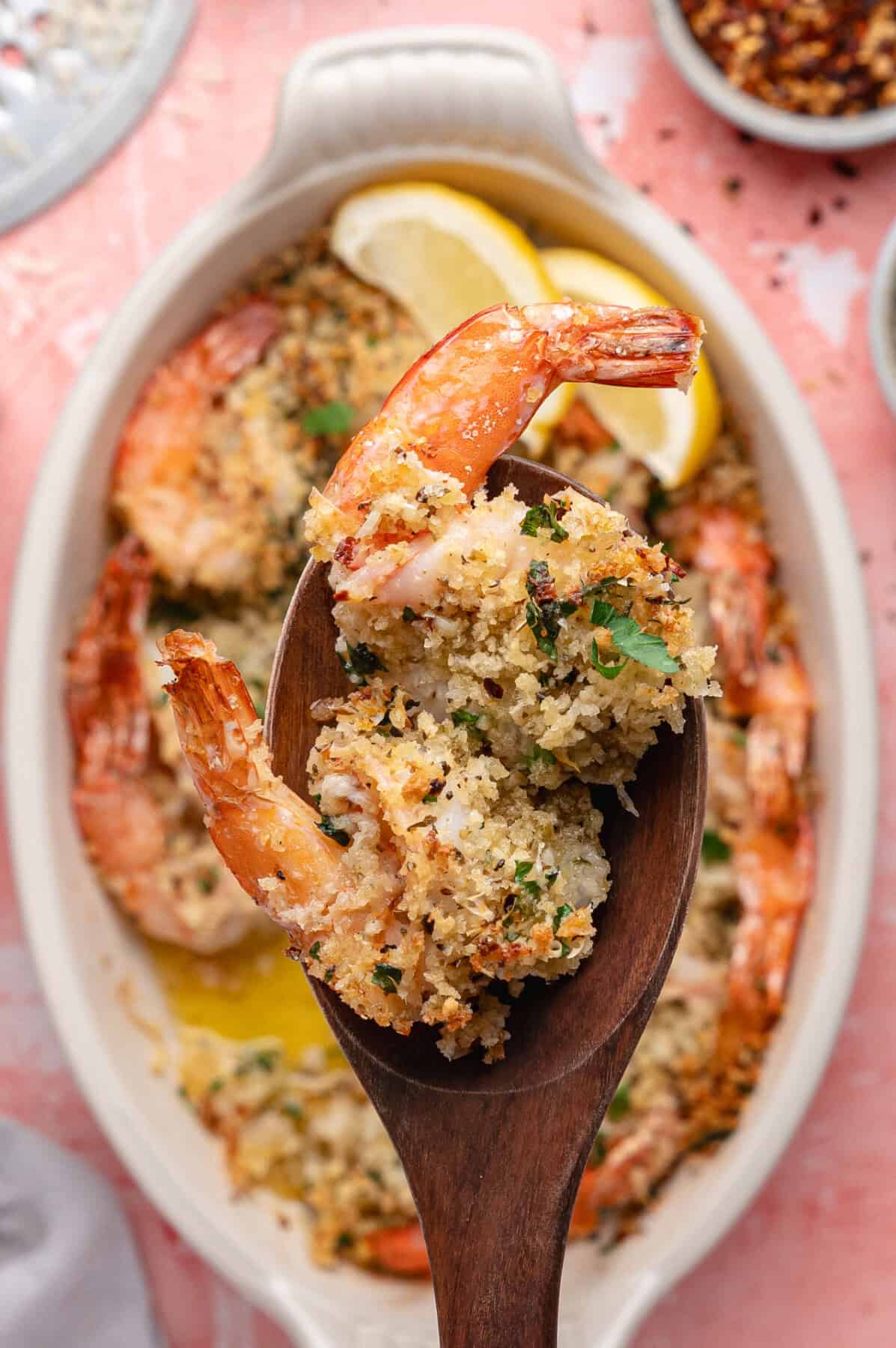 A wooden spoon holds baked shrimp oreganata topped with golden breadcrumbs and herbs, over a white dish filled with more shrimp and two lemon wedges.