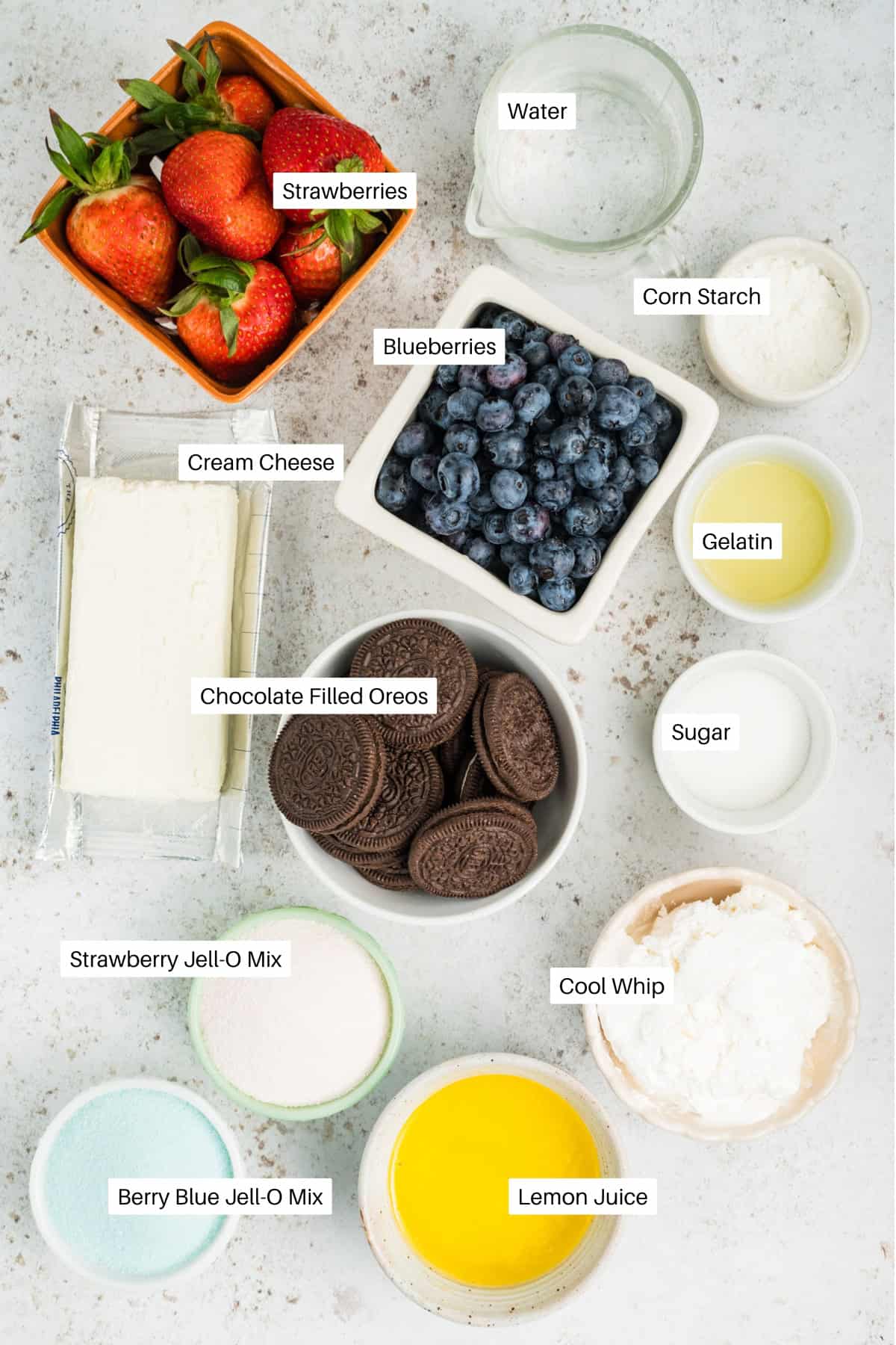 A flat lay of ingredients in bowls for a Berry Cake: strawberries, blueberries, cream cheese, chocolate-filled Oreos, sugar, corn starch, gelatin, water, cool whip, lemon juice, strawberry Jell-O mix, and berry blue Jell-O mix.