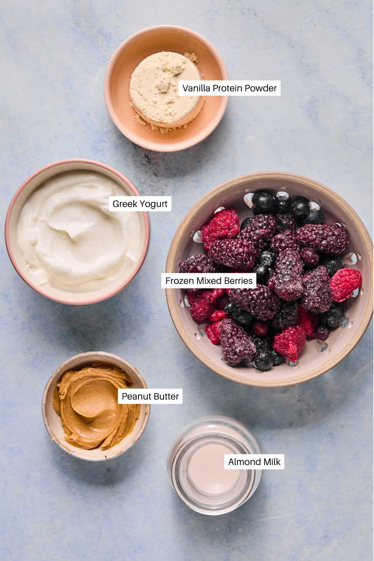 A flat lay of ingredients for a berry smoothie bowl on a light blue surface: vanilla protein powder, Greek yogurt, frozen mixed berries, peanut butter, and almond milk, each neatly labeled with text.
