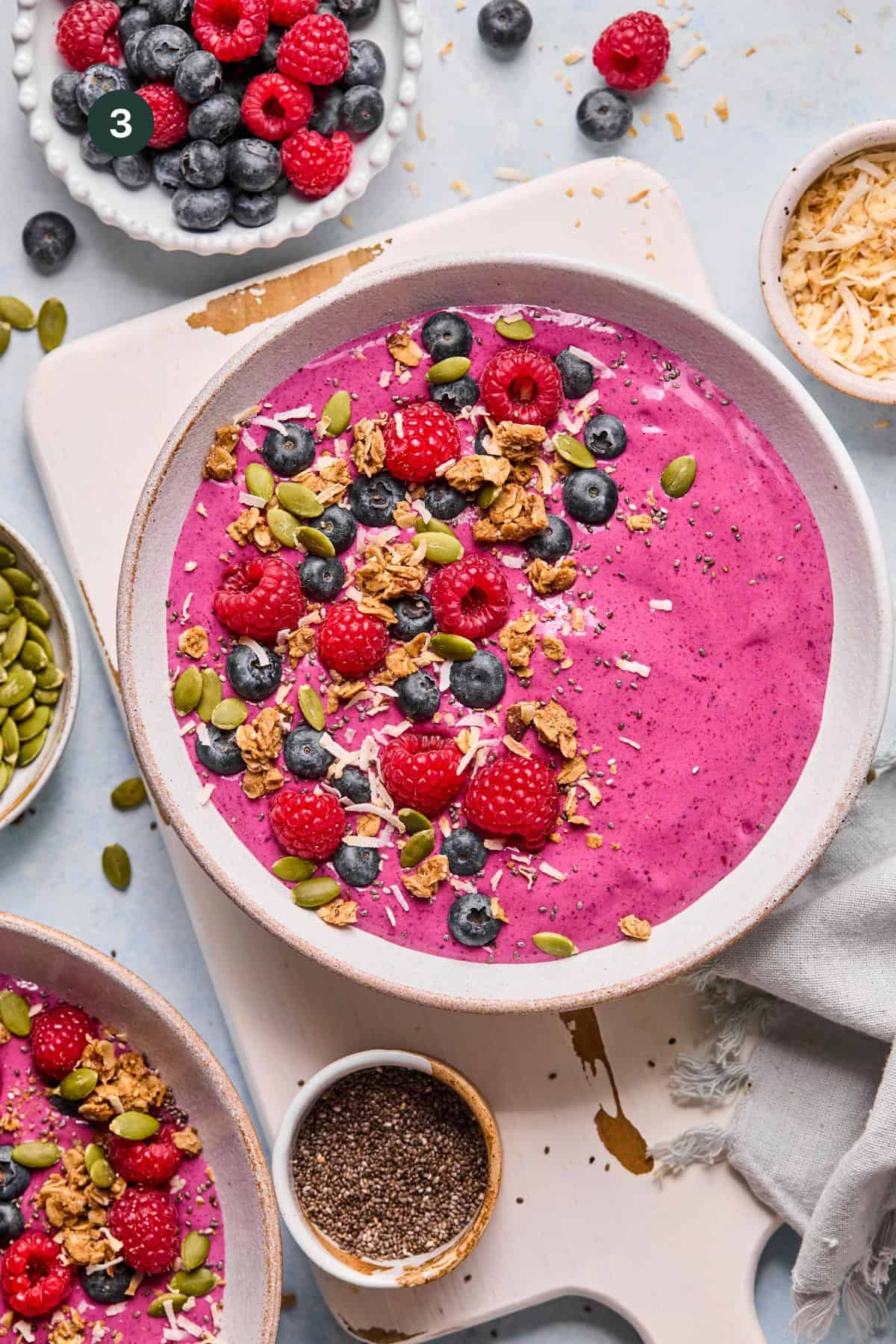 A vibrant berry smoothie bowl topped with raspberries, blueberries, granola, pumpkin seeds, and shredded coconut sits on a white board, surrounded by bowls of berries, seeds, and toasted coconut.