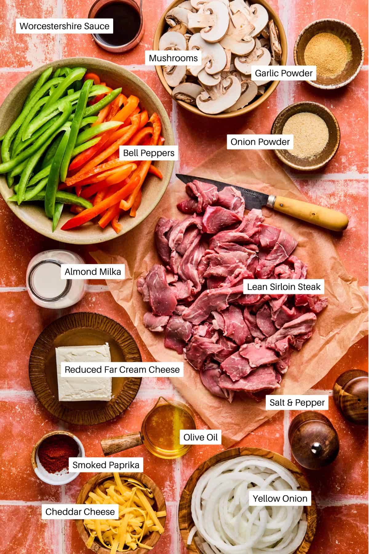 Sliced lean sirloin steak is surrounded by bowls of sliced bell peppers, mushrooms, yellow onion, cheddar cheese, and other Philly cheesesteak bowls essentials on a tiled surface.