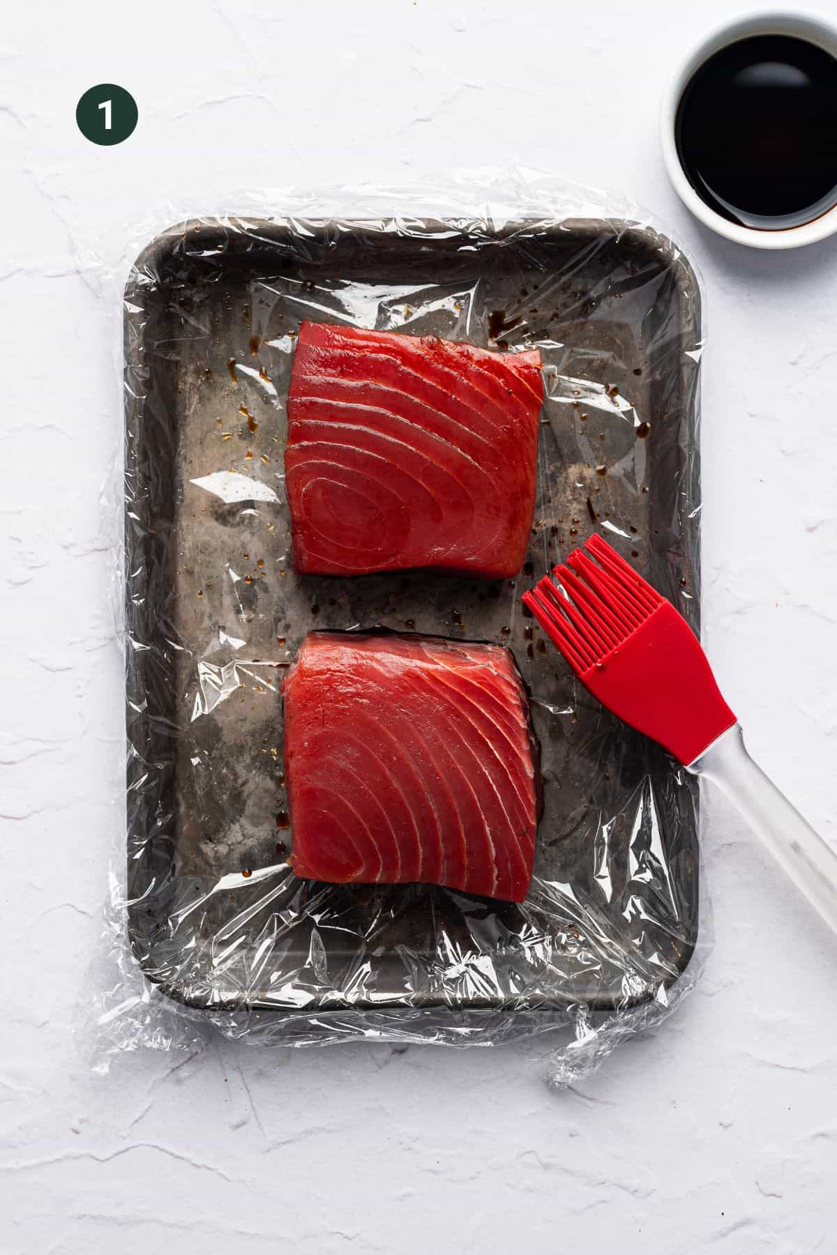 Two raw tuna steaks sit on a plastic-wrapped baking tray next to a red silicone basting brush, while a small bowl of dark marinade or soy sauce rests in the upper right cornerโideal prep tools for tuna or baked chicken cutlets alike.